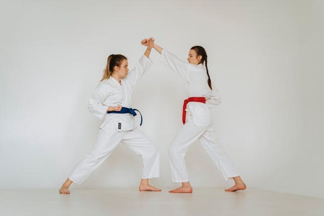 Dwie dziewczyny w karate-gi jedna z niebieskim, a druga z czerwonym pasem. Jedna wykonuje blok ko age uke, a druga uderza oi tsuki jodan. Obie są w pozycji zenkutsu dachi.